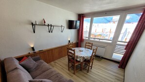 Una sala de estar acogedora con un sofá marrón, una mesa de comedor de madera con cuatro sillas, un televisor montado en la pared y una ventana que ofrece vistas de un paisaje nevado.