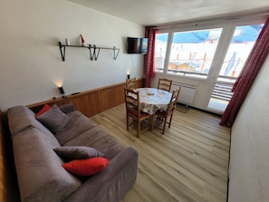 A cozy living room with a brown sofa, a wooden dining table with four chairs, a mounted TV, and a window with a view of a snowy landscape.
