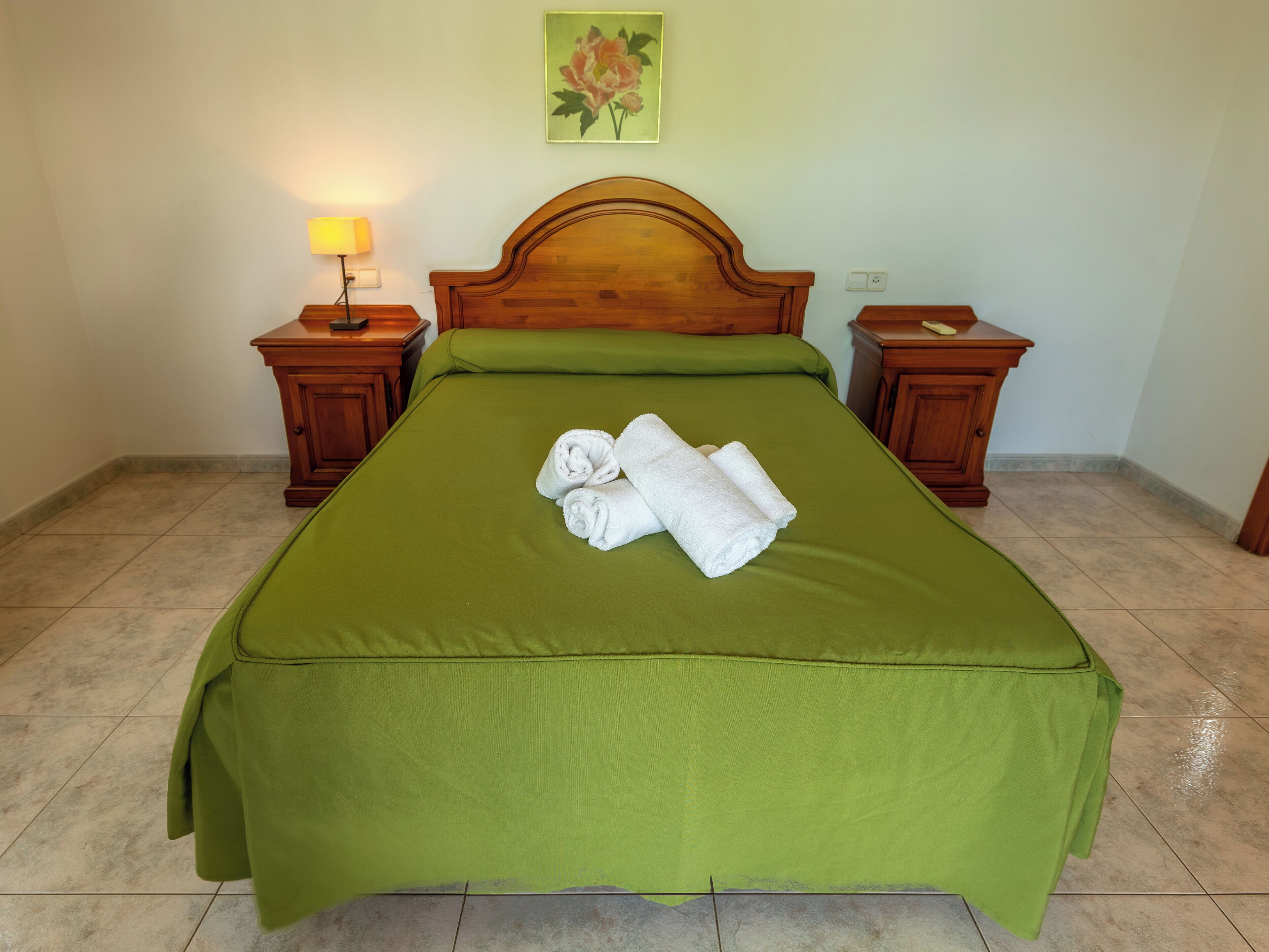A neatly made bed with green bedding and rolled white towels, wooden headboard, two bedside tables with lamps, and a floral painting on the wall.