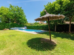 Piscina cercada por gramado, guarda-sol de palha e árvores verdes exuberantes.