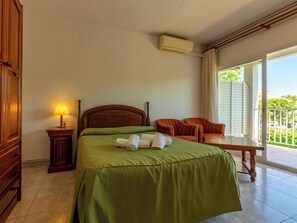 Quarto de hotel com cama, mesa de cabeceira com luminária, duas poltronas listradas, uma mesinha e uma varanda com vista.