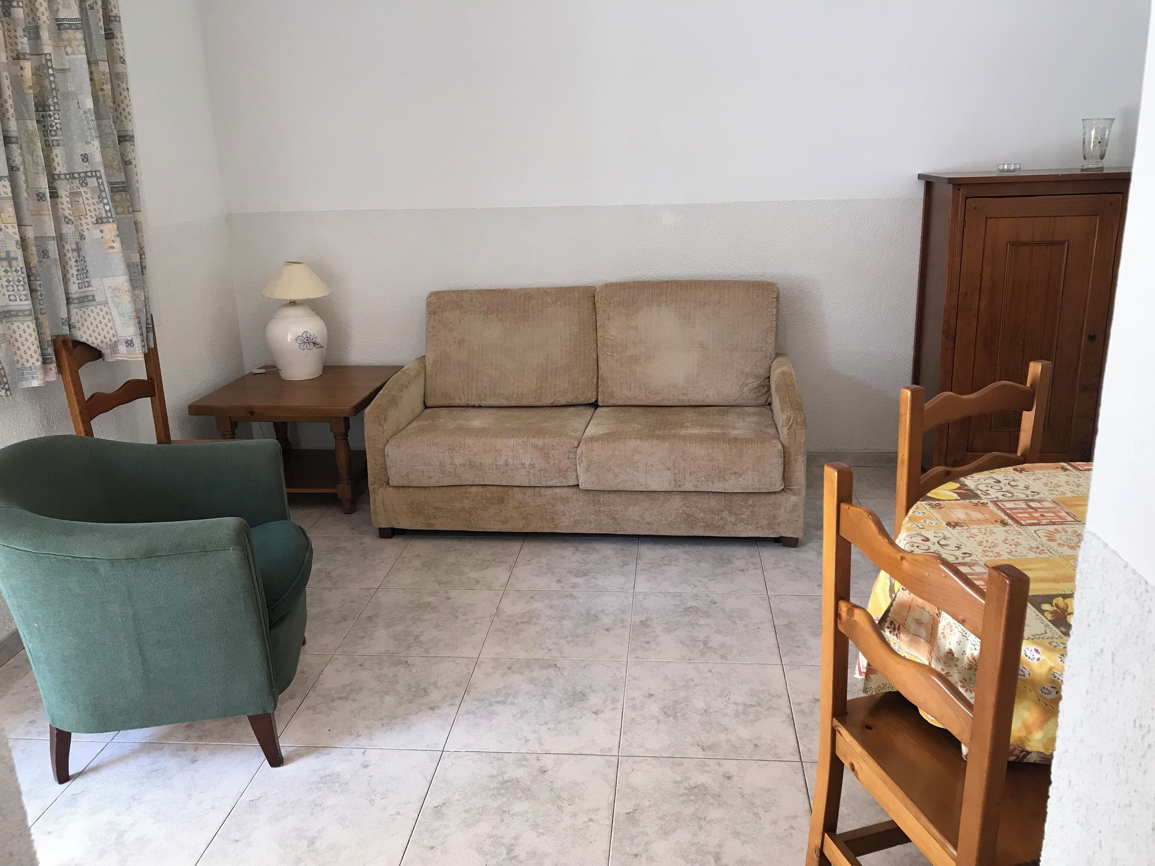 A small, furnished room with a sofa, a green armchair, a wooden table with a lamp, and a wooden cabinet.