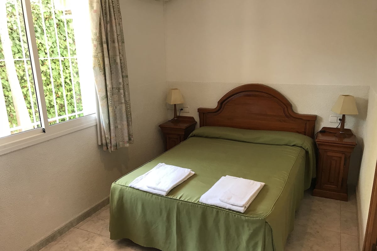 Ein Zimmer mit einem Bett, zwei Nachttischen, einem Fenster mit Vorhängen und Blick ins Grüne.