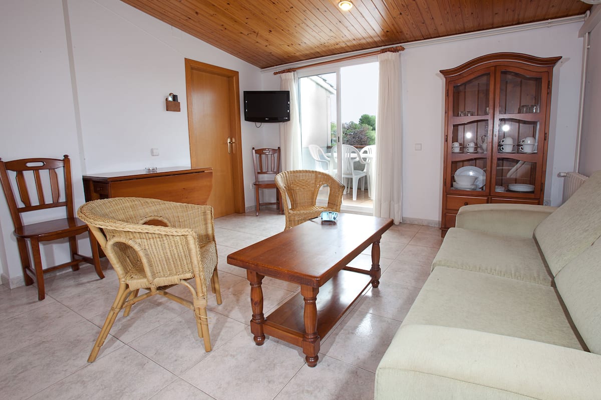 Ein Wohnzimmer mit Holzboden, einer Couch, einem Tisch, Korbstühlen, einer Vitrine mit Glastüren und einem Fernseher an der Wand.