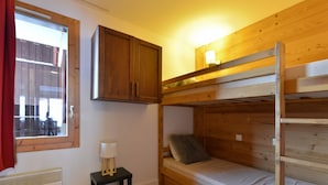 Een slaapkamer met stapelbed, een raam, een verwarming, een klein tafeltje met lamp en houten wanden.