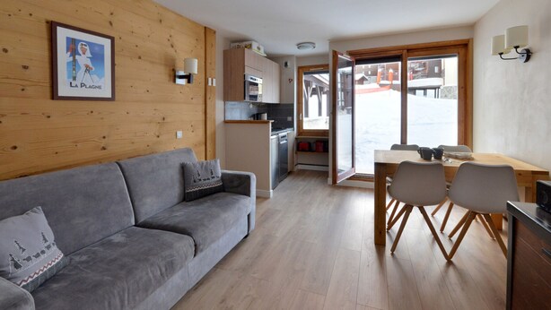 Een gezellige woonkamer met een grijze bank en een eettafel met stoelen. In de achtergrond is een keuken met een fornuis en kastjes te zien.