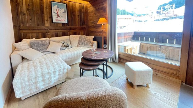 Ein gemütliches Zimmer mit Holzbalkendecke, einer Sofagarnitur, einem runden Tisch mit zwei Gläsern und Blick auf einen Balkon mit Sitzgelegenheiten.