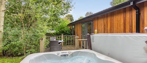 Een hottub met een houten terras, een houten gebouw met een glazen deur, en een omheind gebied met begroeiing.