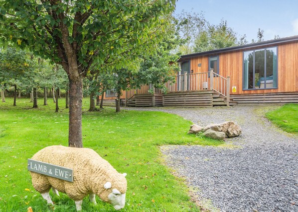 A wooden cabin with a gravel path leading to a wooden deck, surrounded by greenery and a sheep labeled 'LAMB & EWE'.