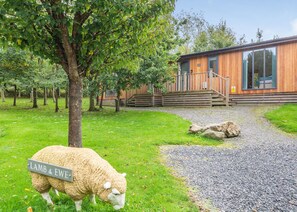 Une cabane en bois avec un chemin de gravier menant à une terrasse en bois, entourée de verdure et d’un mouton portant une étiquette indiquant « AGNEAU ET BREBIS ».