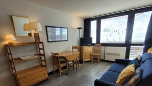 Ett kompakt vardagsrum med en soffa, ett litet bord, ett skrivbord och en stol. På skrivbordet står en tv, på bordet står en lampa och på väggen hänger en inramad tavla.