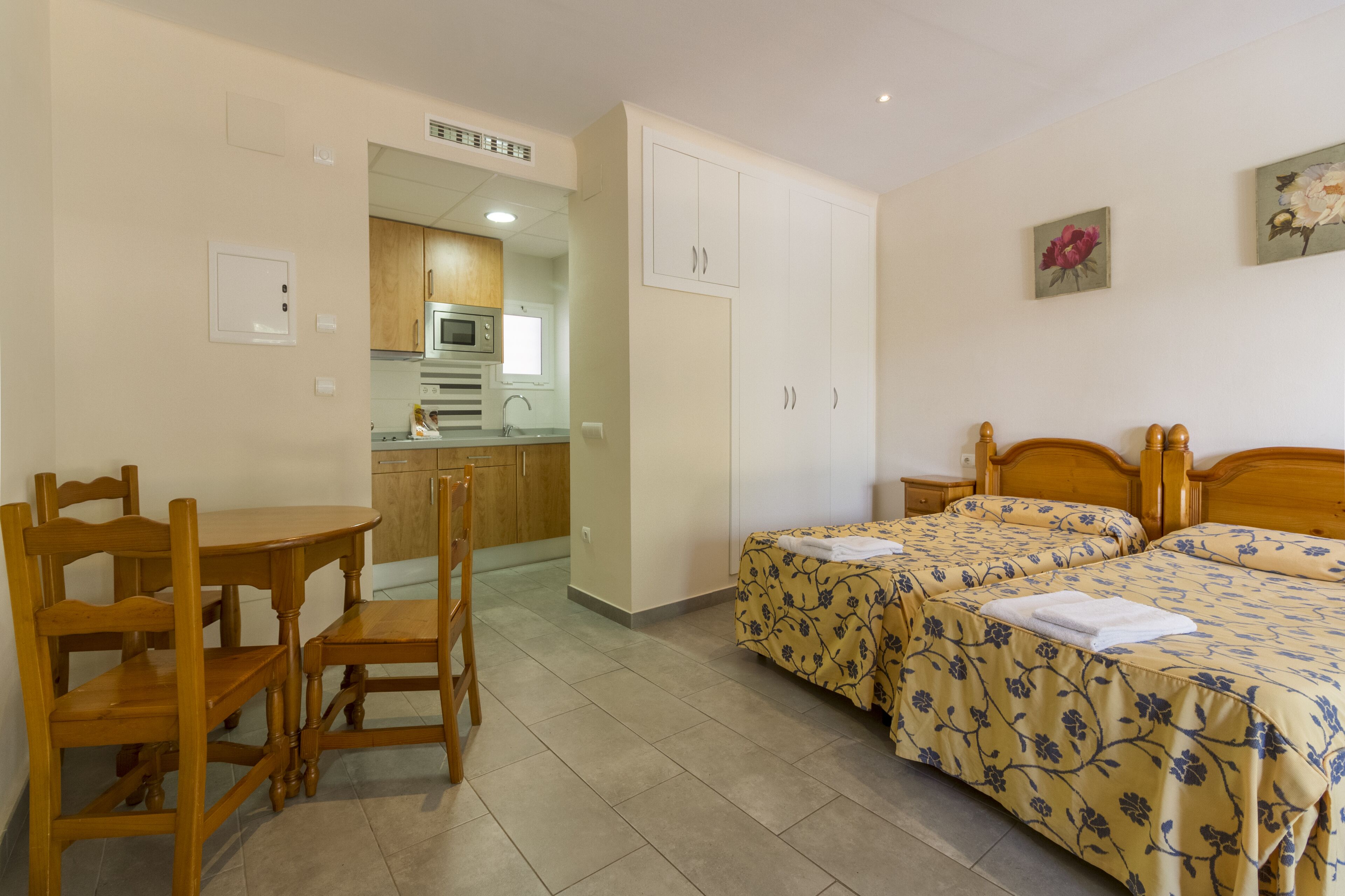 A bedroom with two beds, a wooden desk, and a kitchenette in the background.