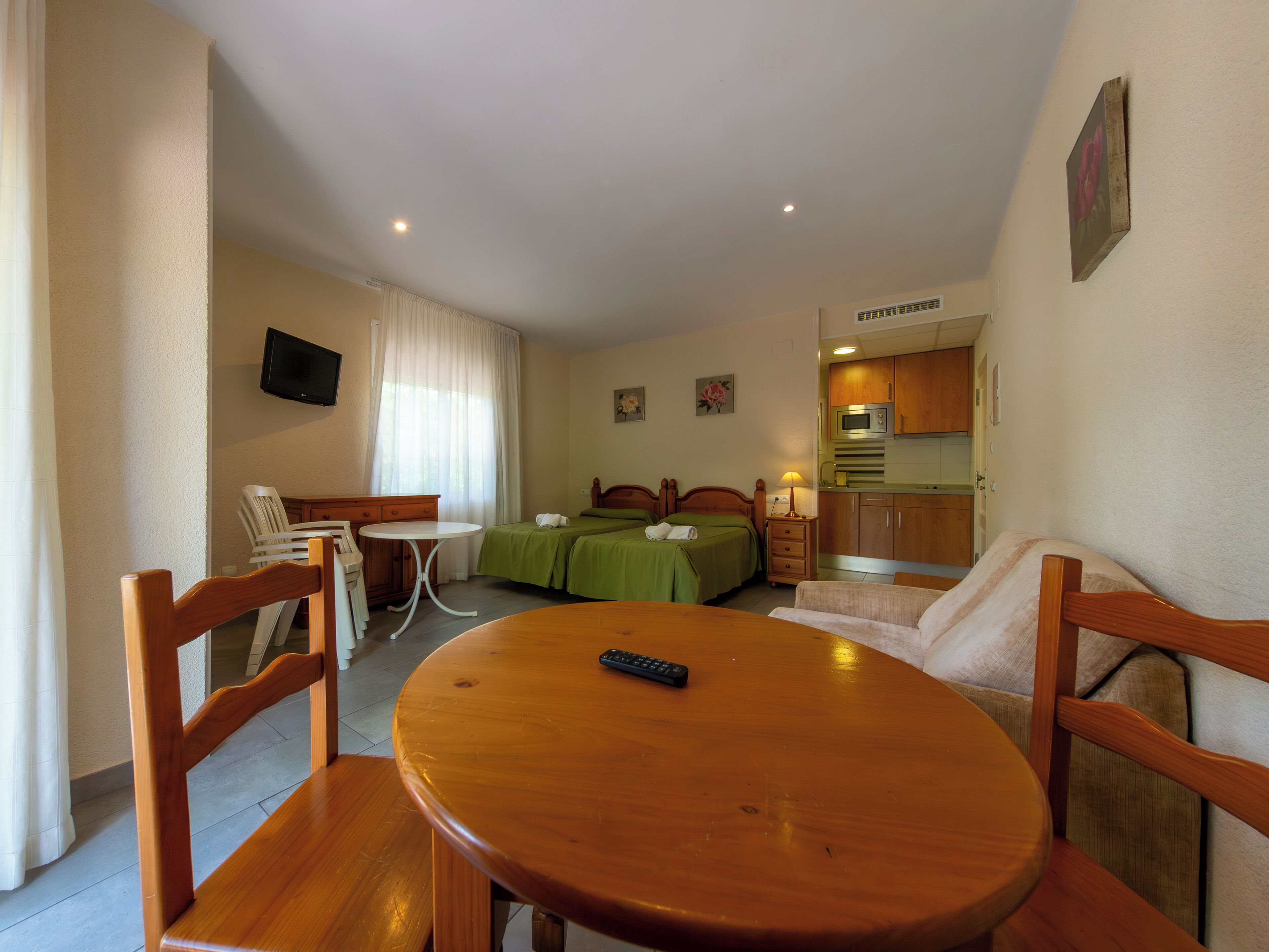 Habitación de hotel con mesa de comedor de madera, dos camas individuales, kitchenette, televisor montado en la pared y ventana con cortinas.