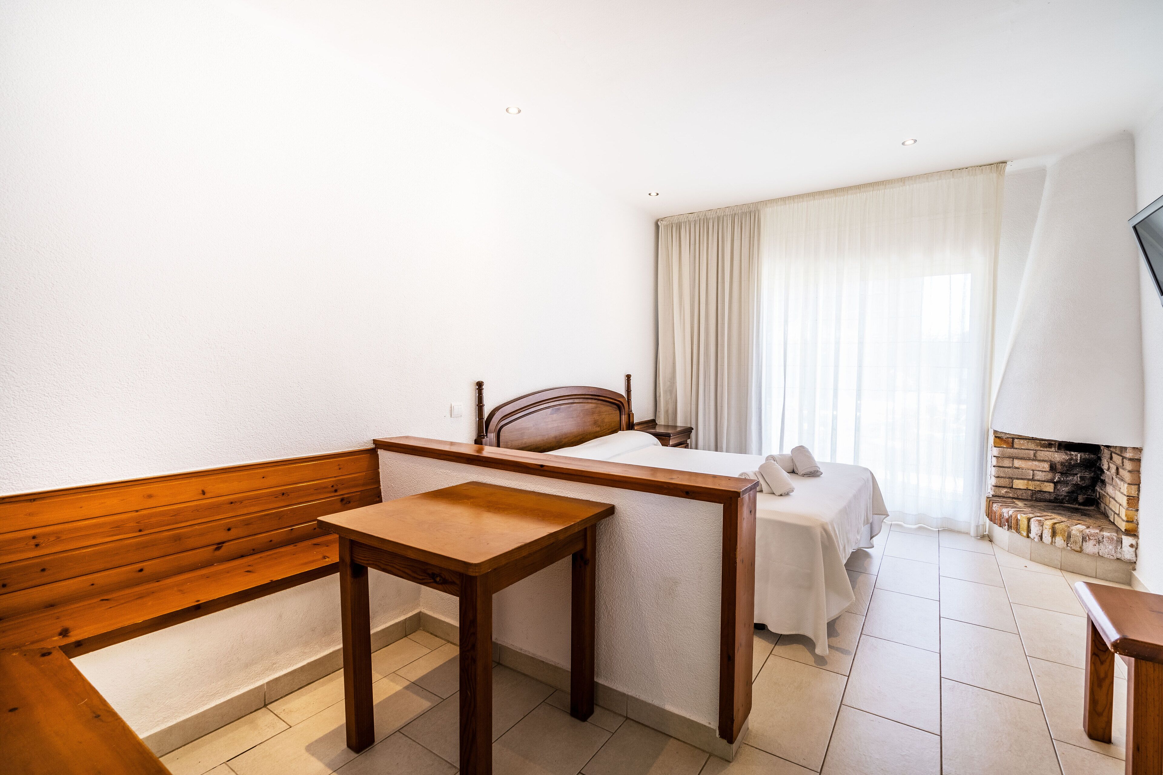 A hotel room with a wooden bench, a small table, a bed with a white bedspread, a television, and a fireplace.