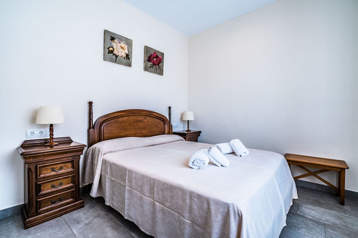 Una cama bien hecha con cabecera de madera, una mesita de noche de madera con una lámpara y un pequeño banco de madera.