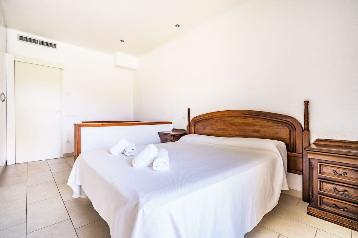 Una cama bien hecha con sábanas y toallas blancas, un cabecero de madera, una mesita de noche con una lámpara y un aparador de madera con cajones.