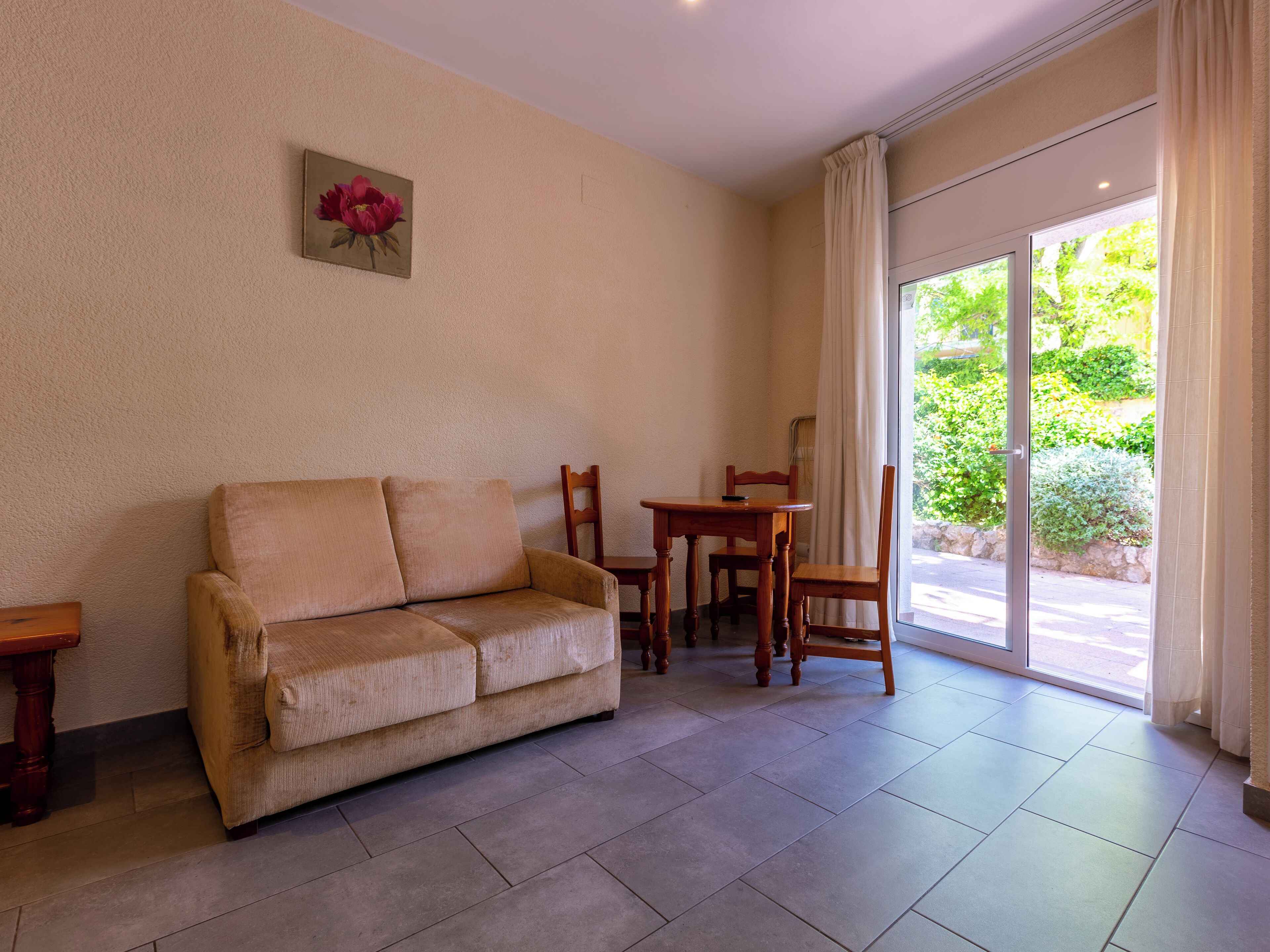 Un salon avec un canapé beige, une table et des chaises en bois, un tableau au mur et, par une porte vitrée, une vue sur la verdure.