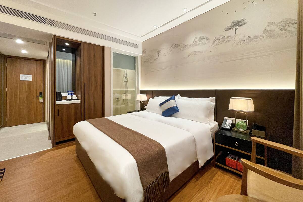 Ein Hotelzimmer mit einem großen Bett, einem Holzschrank, einer Glasdusche, einem Nachttisch mit Lampe und einer Wand mit Wandgemälde.