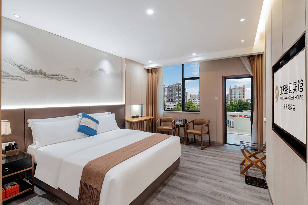 Ein Hotelzimmer mit einem großen Bett, einem Schreibtisch mit Stuhl, einem kleinen Tisch für zwei Personen und Blick auf Gebäude durch ein Fenster.