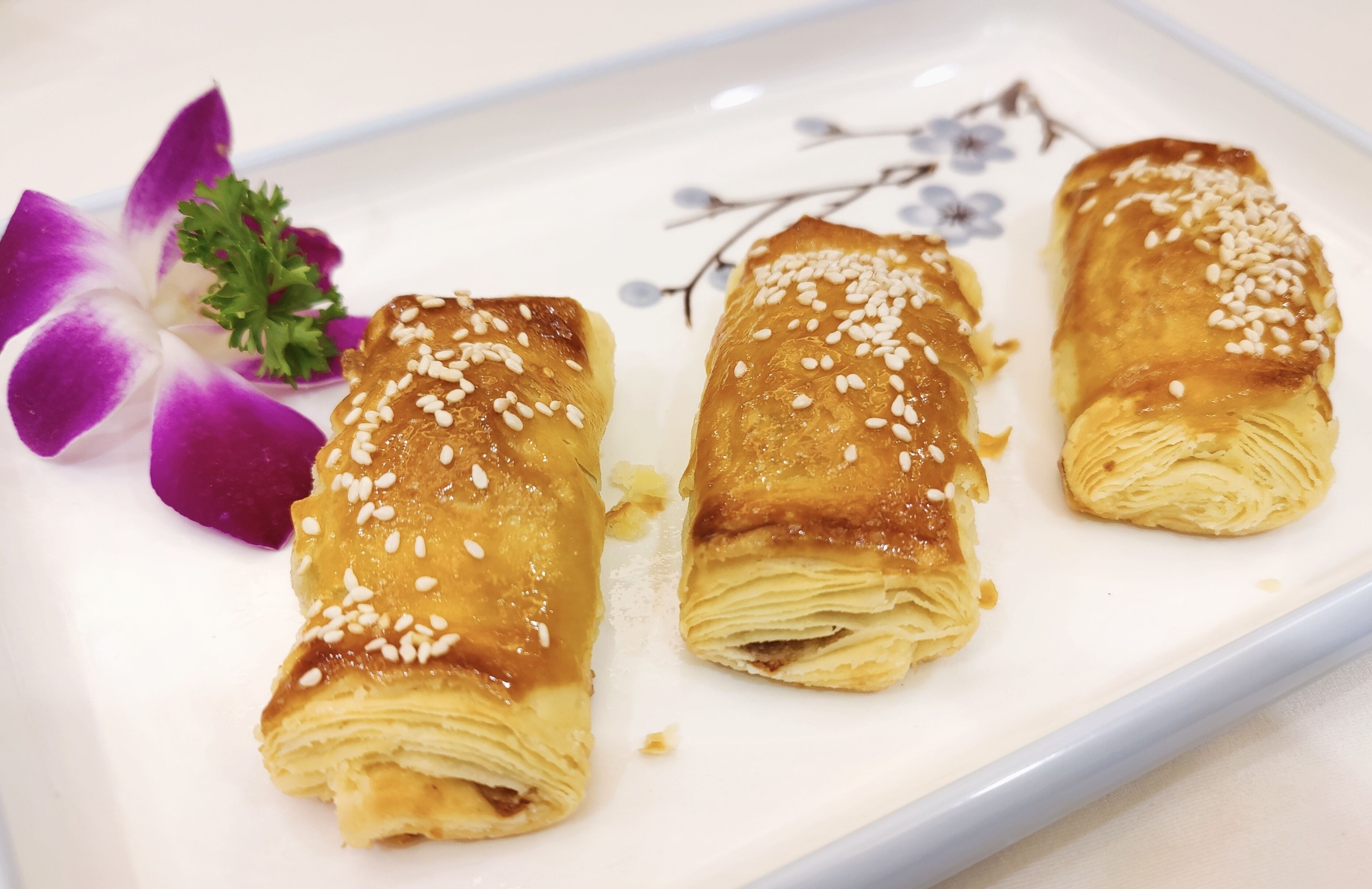 Three pastries on a white plate with sesame seeds, accompanied by a purple orchid and a sprig of parsley.