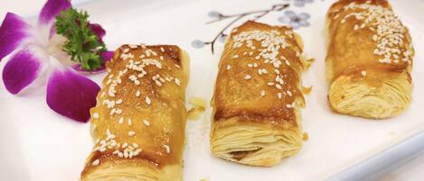 Three pastries on a white plate with sesame seeds, accompanied by a purple orchid and a sprig of parsley.
