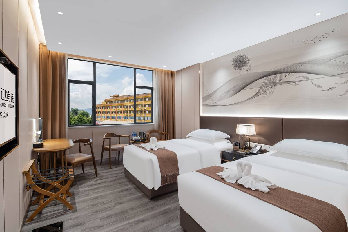 Ein Hotelzimmer mit zwei Betten, einem Schreibtisch, Stühlen, einem großen Fenster mit Blick auf ein Gebäude und einem Wandbild.
