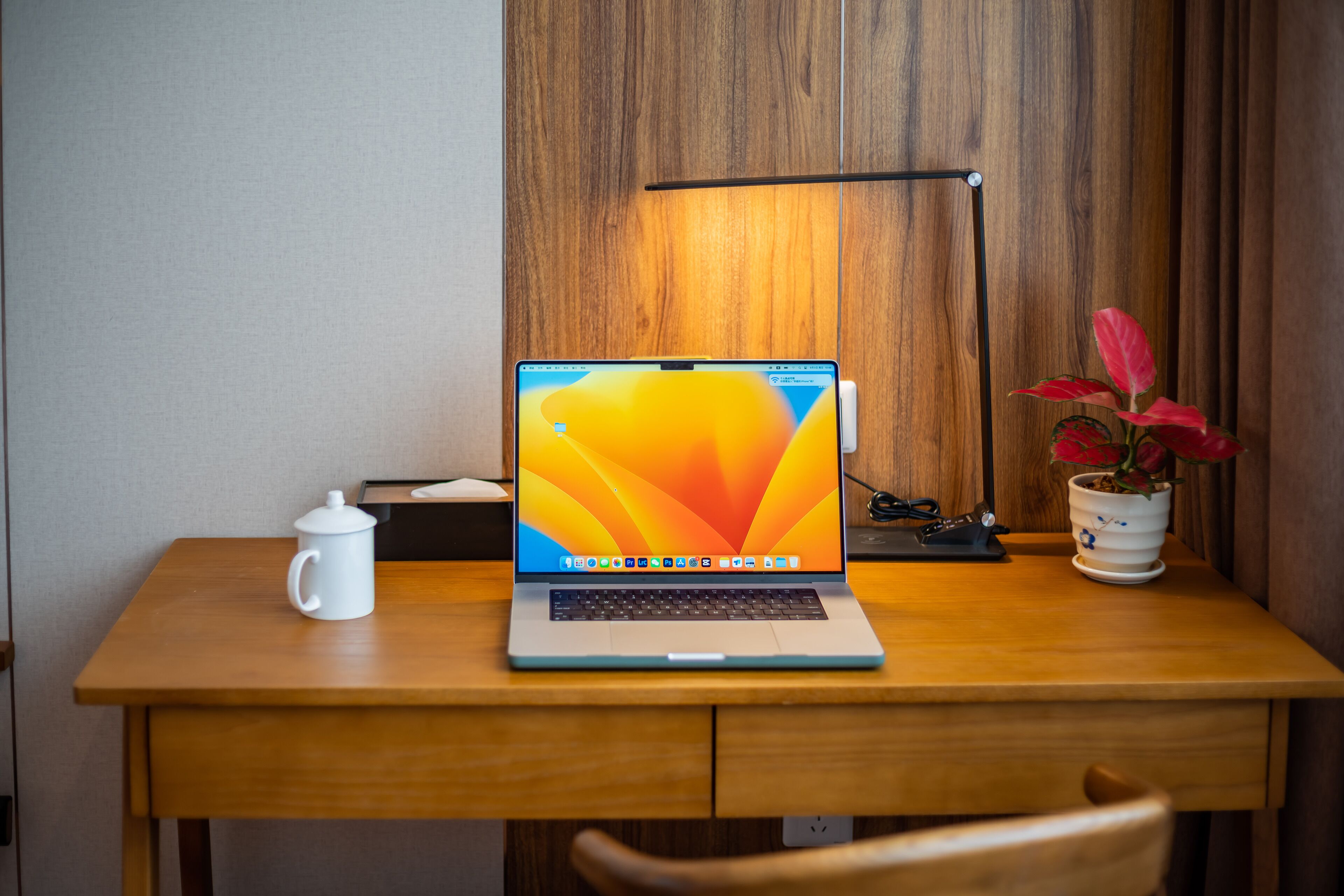 A wooden desk with a laptop, a white mug, a lamp, and a potted plant.