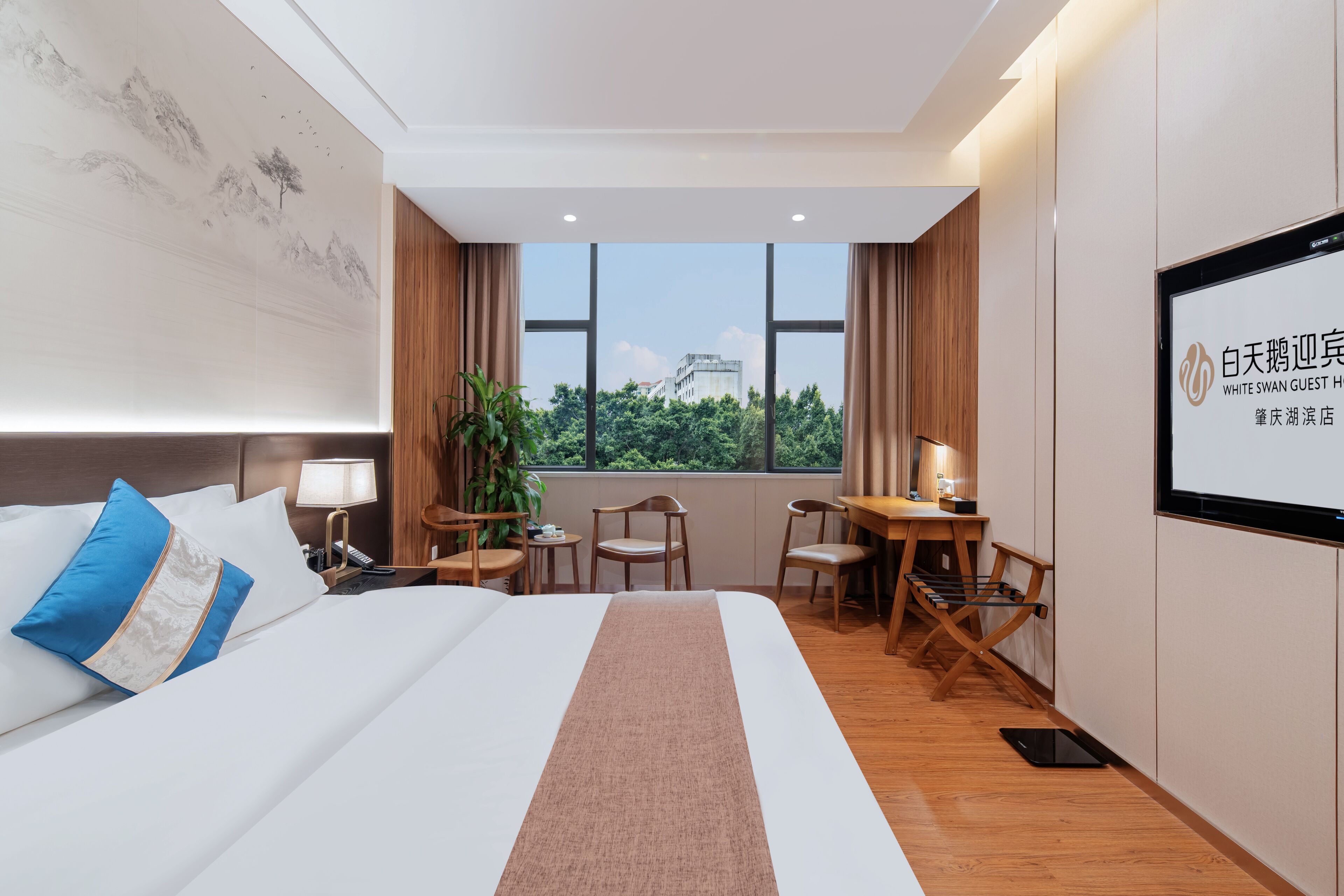 A hotel room with a bed, a desk, a chair, a TV, and a window with a view of trees and buildings.