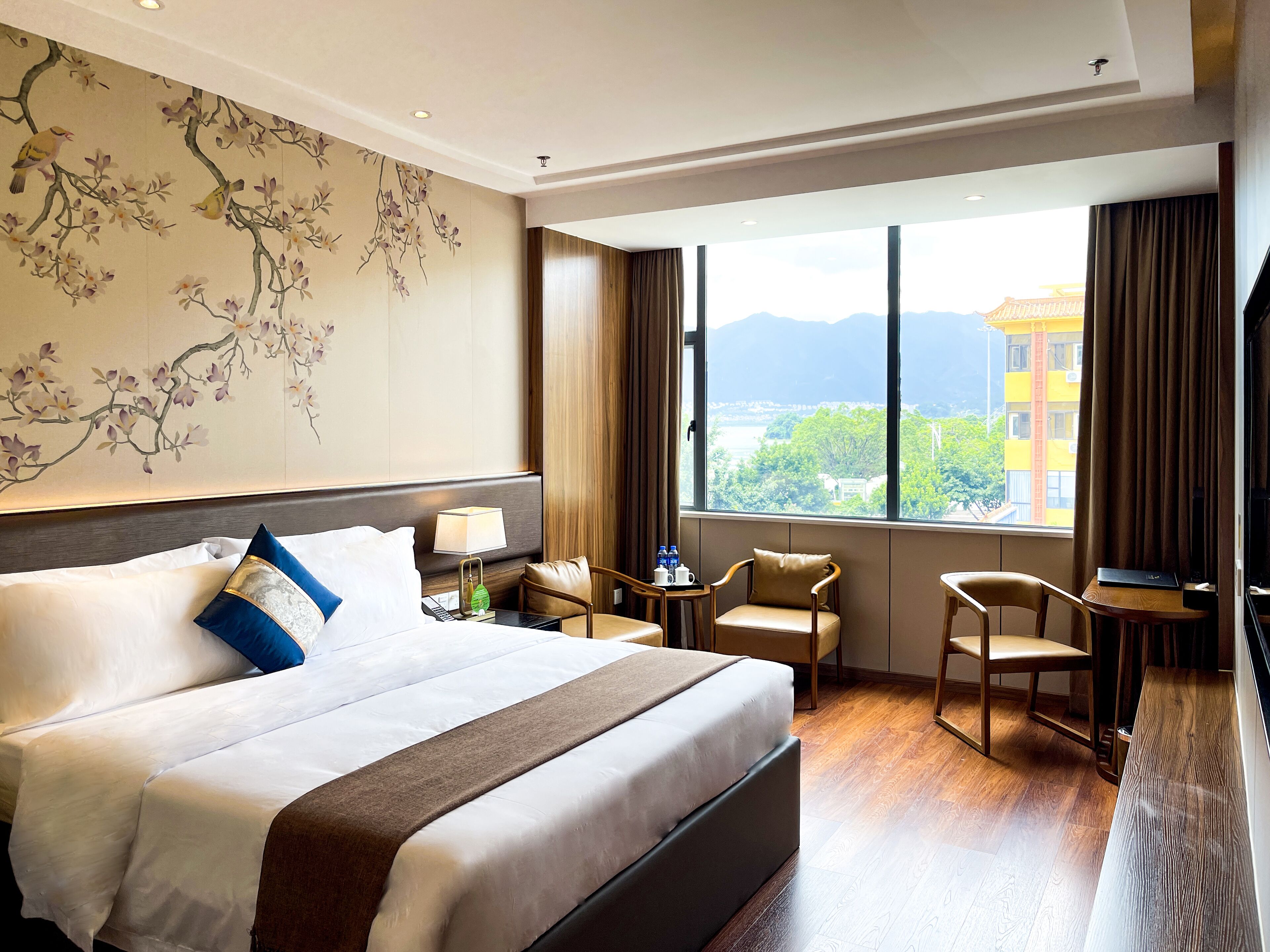 Habitación de hotel con una cama grande, un escritorio, dos sillas, una lámpara y vistas a un paisaje urbano a través de amplios ventanales.