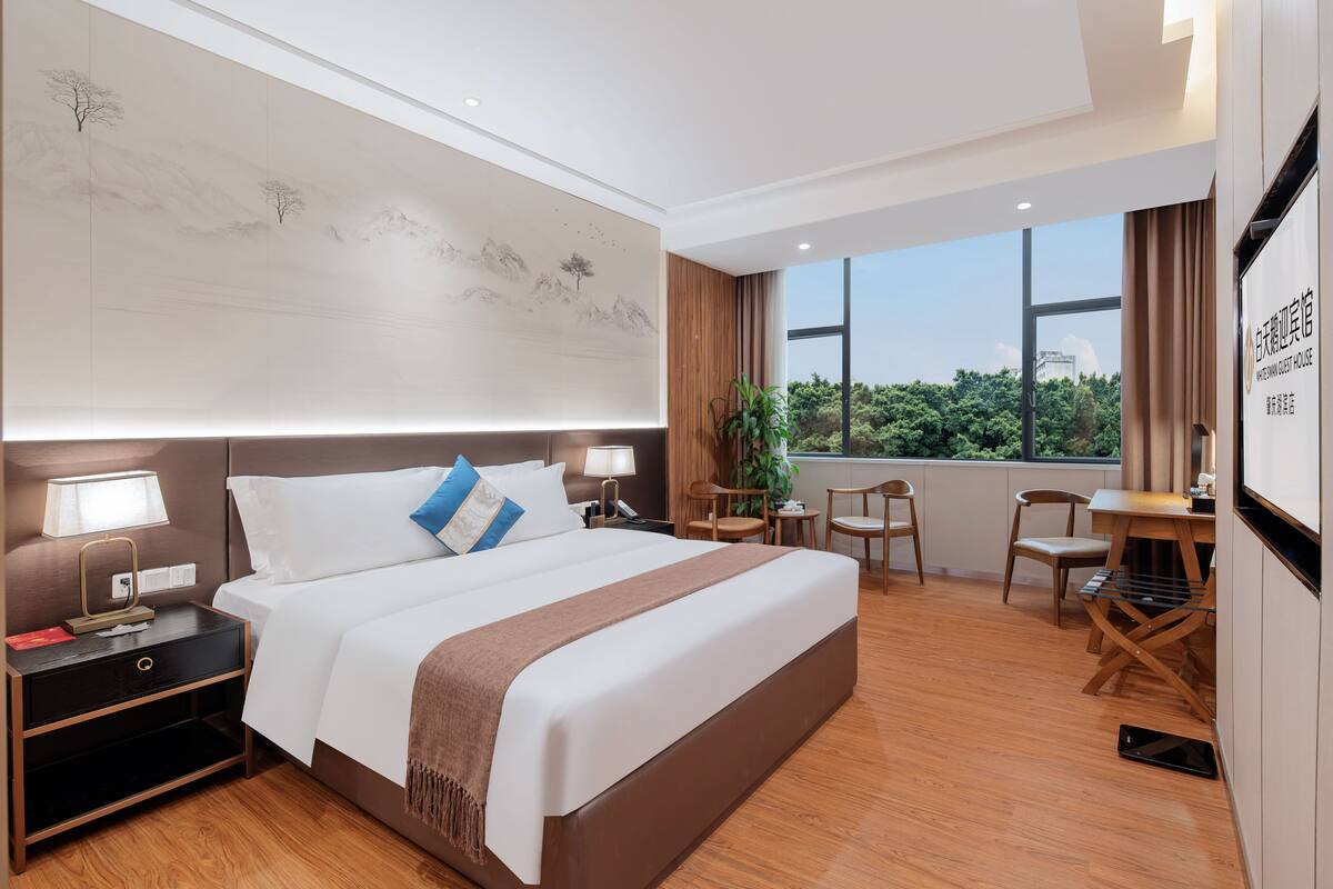 Une chambre d’hôtel avec un grand lit, des tables de chevet, un bureau et une chaise. Il y a une fenêtre donnant sur un paysage verdoyant.