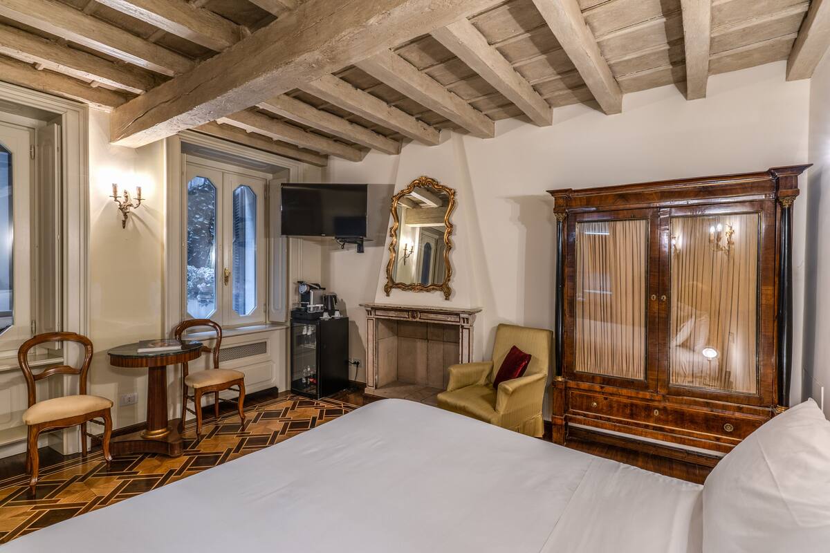 A hotel room with a large bed, wooden ceiling beams, a fireplace, a TV, and a large mirror.
