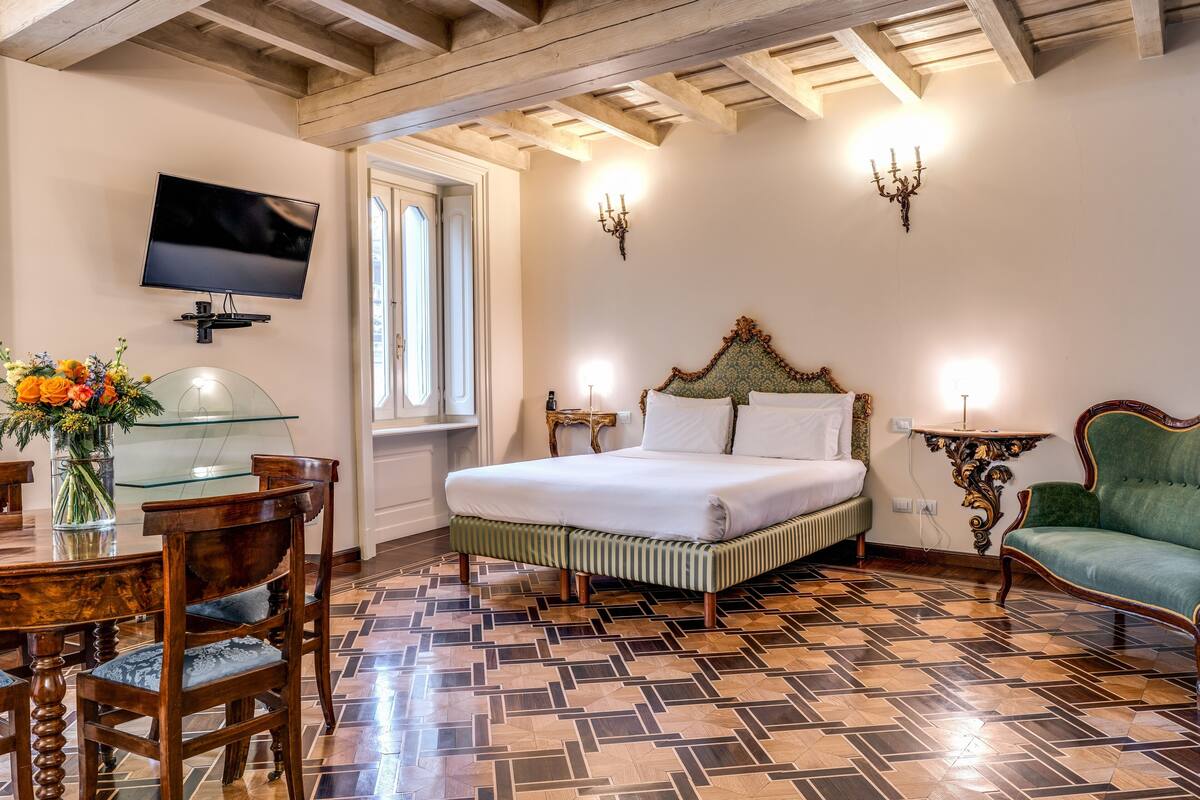 A bedroom with a bed, a TV mounted on the wall, a dining table with chairs, and a chandelier.