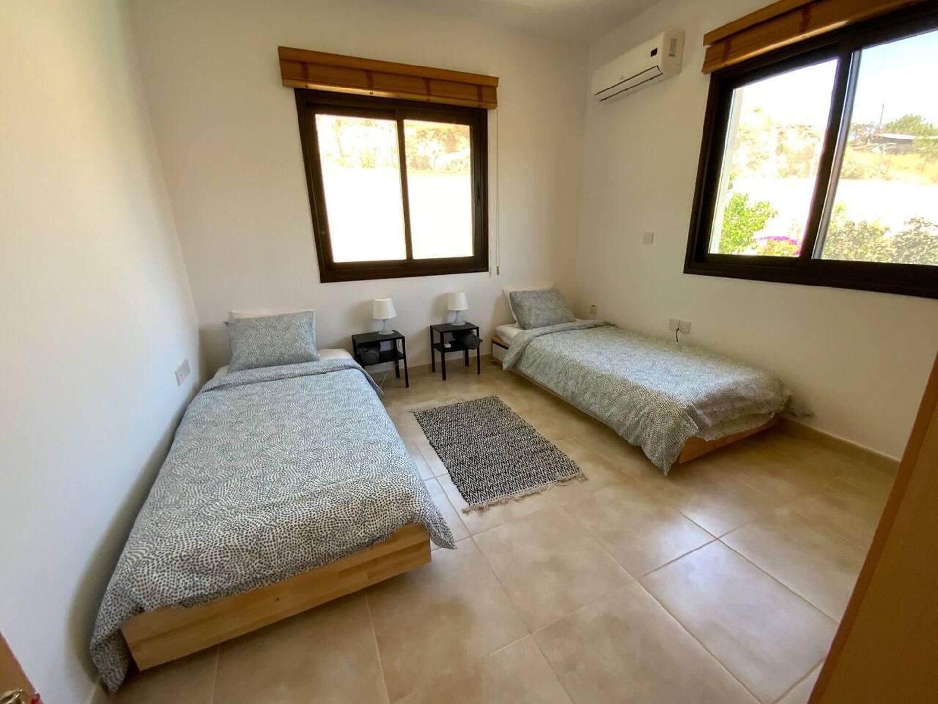 A small, clean room with two single beds, a small table, two bedside lamps, and a window providing natural light.