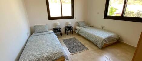 A small, clean room with two single beds, a small table, two bedside lamps, and a window providing natural light.