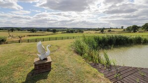 A serene landscape with a swan statue on a brick pedestal by a pond, surrounded by grassy fields and a wooden fence. - Far Coley Farm &amp; Log Cabin (Stafford)