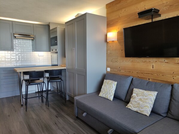 A compact living space with a kitchenette, a sofa with patterned cushions, and a mounted TV.