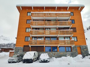 Un immeuble à plusieurs étages doté de balcons, d’une base en pierre et de voitures garées recouvertes de neige.