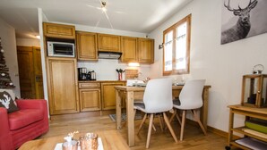 Et kompakt køkken med træskabe, et spiseområde med to stole, et lille bord med stearinlys og et billede af et rådyr på væggen.