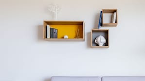 Une pièce au décor minimaliste, avec un mur blanc, un canapé de couleur violet pâle et trois étagères en bois ornées de livres et d’objets décoratifs.