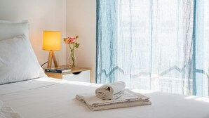 Un lit bien fait, avec du linge de lit blanc, une lampe de chevet, un livre et une serviette pliée sur une table de chevet.
