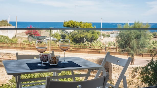 Une table avec deux verres de vin et un bol de collations, placée sur un balcon donnant sur un paysage côtier.