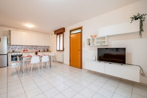 Una cocina moderna con armarios blancos, un comedor con sillas, un televisor sobre un mueble blanco y una puerta con ventanal.