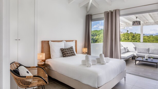 A bedroom with a large bed, a wicker chair, a sofa, and a view of greenery outside.