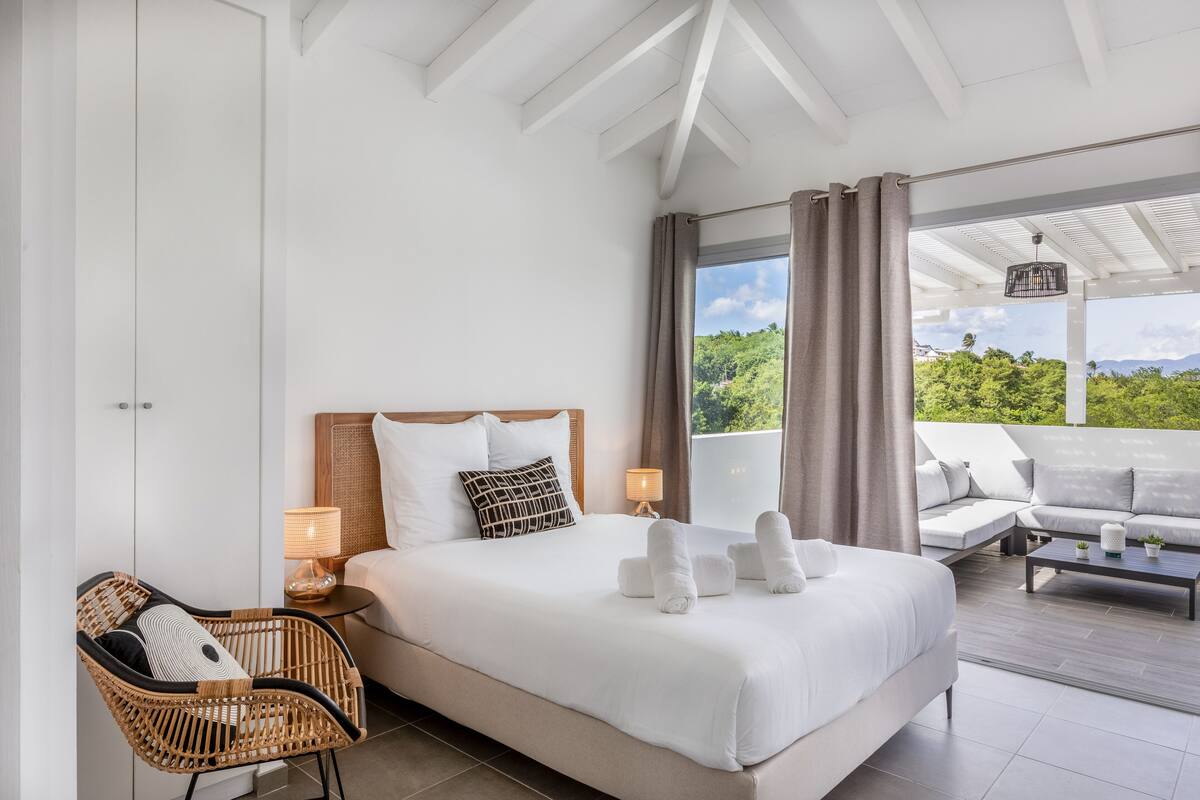 Une chambre avec un grand lit, un fauteuil en osier, un canapé et une vue sur la verdure environnante.