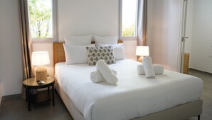 A neatly made bed with white linens and two rolled towels, a bedside lamp, and a small round table with a remote control.