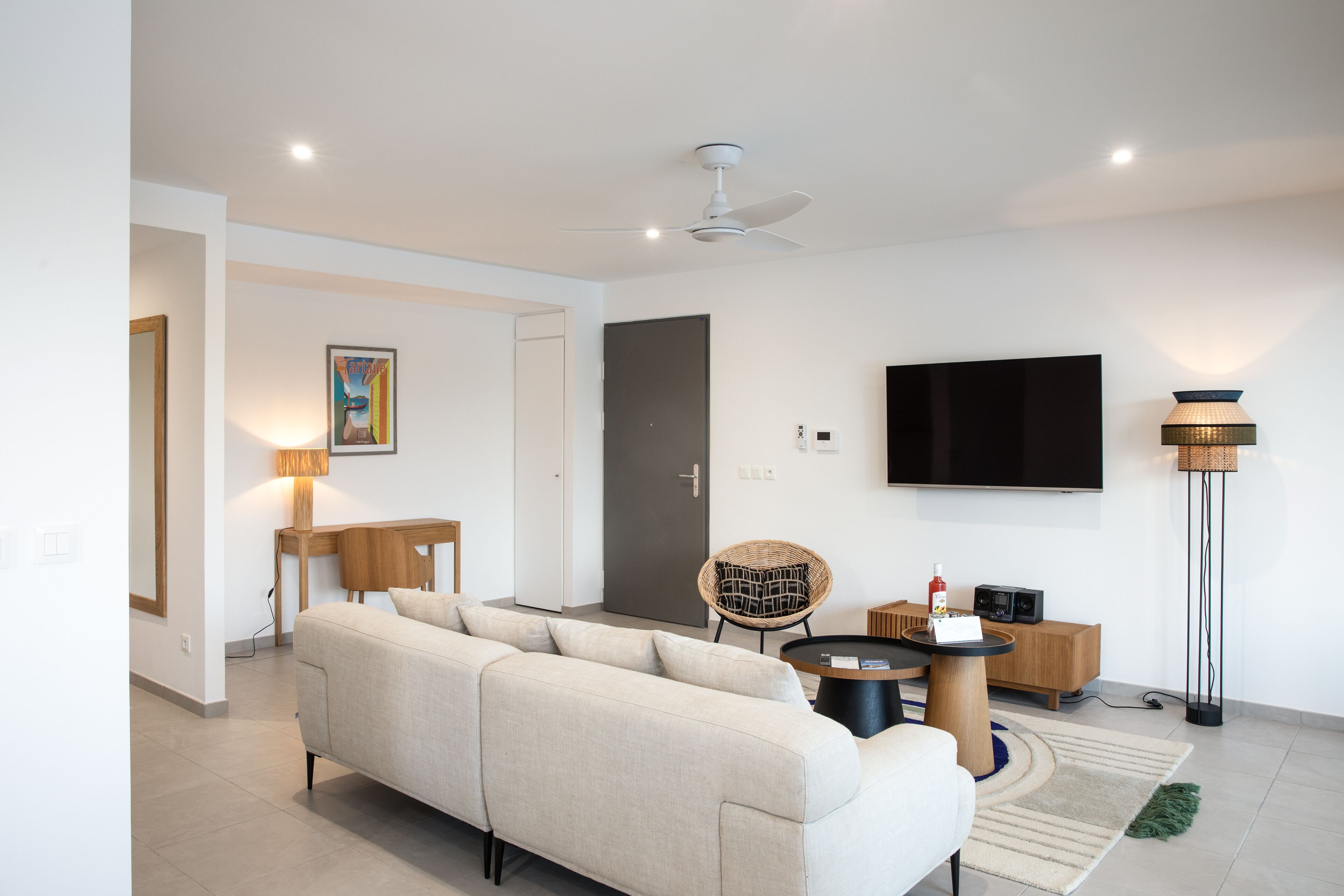 Una sala moderna con un sofá, una mesa redonda de centro, una silla de mimbre, un televisor montado en la pared y una lámpara de pie.