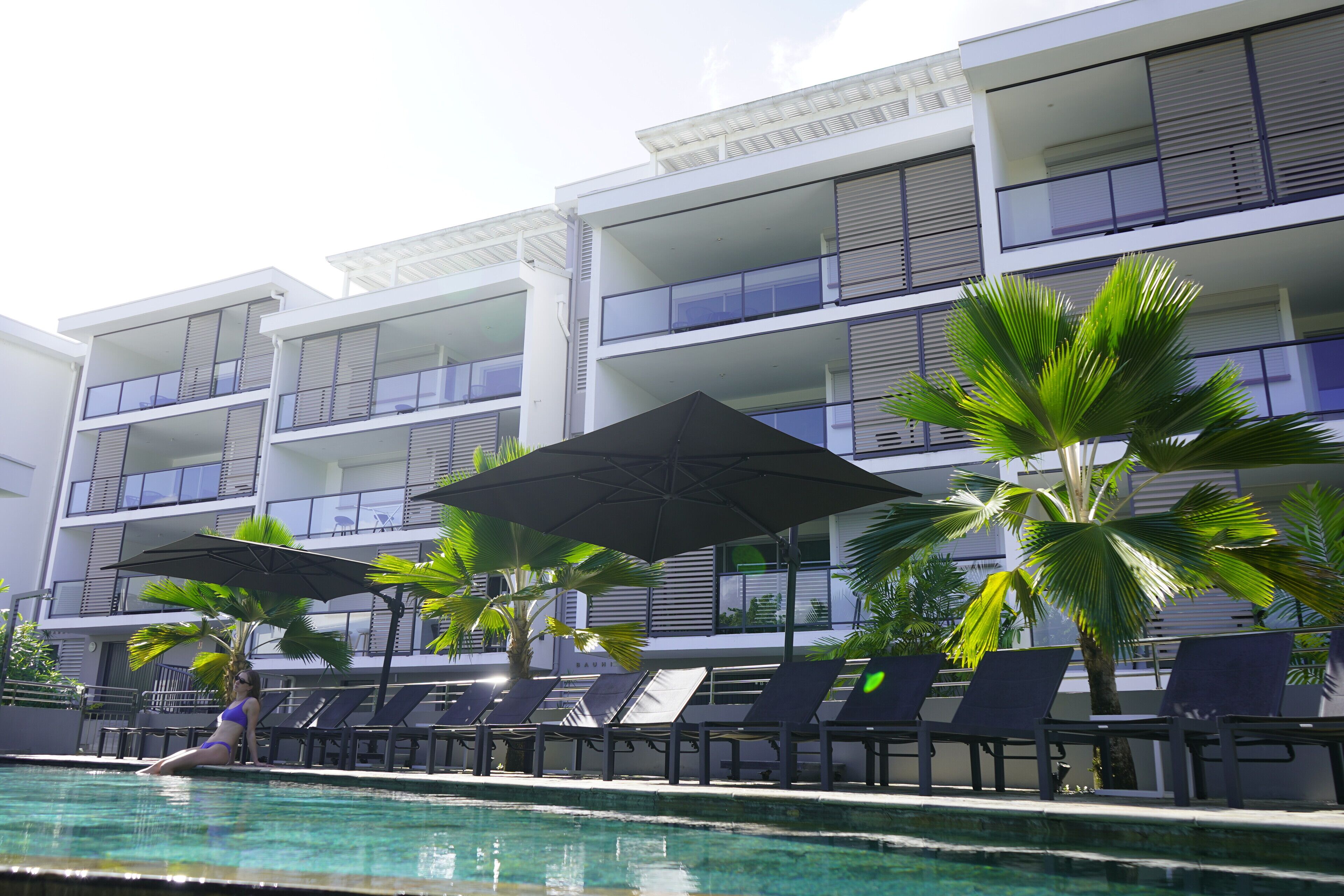 Un espace piscine avec des chaises longues et des parasols, une personne en maillot de bain et un immeuble à plusieurs étages doté de balcons.