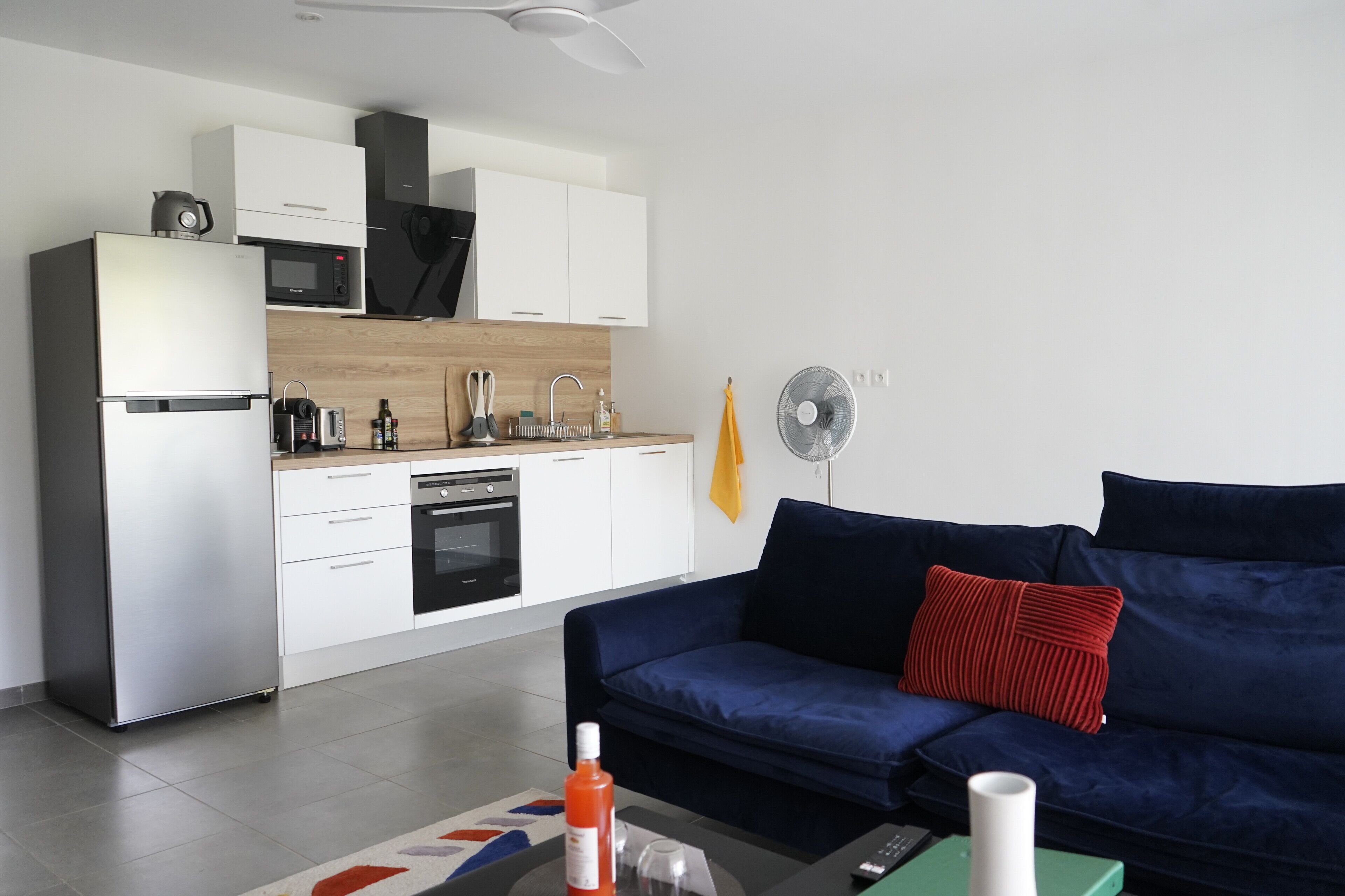A modern kitchen with white cabinets, a microwave, and a dishwasher. There is a blue sofa with a red pillow and a yellow towel hanging on the wall.