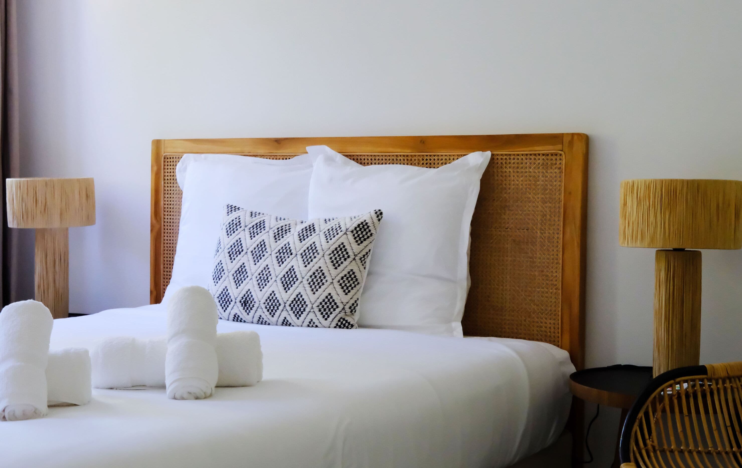 Un lit bien fait, avec du linge de lit blanc et un coussin à motifs, flanqué de deux tables de chevet en bois sur lesquelles sont posées des lampes.