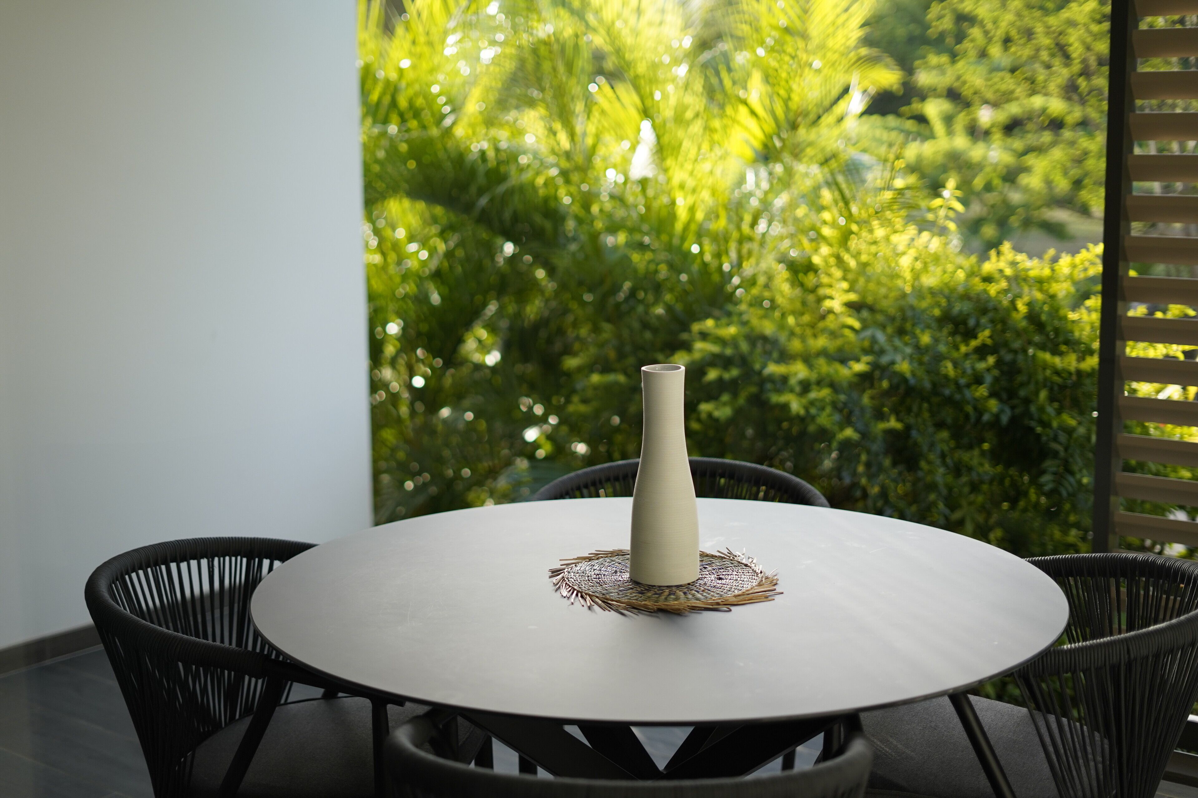 Een ronde tafel met een witte vaas en een geweven placemat, omringd door zwarte stoelen, voor een achtergrond van weelderige groene begroeiing.